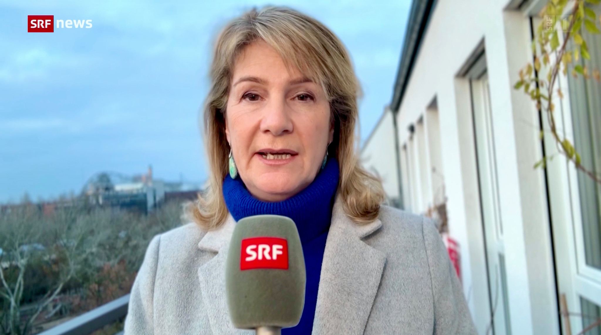 SRF-Deutschland-Korrespondentin Alexandra Gubser in grauem Mantel mit SRF-Mikrofon, im Freien auf einem Balkon, blauer Rollkragenpullover. Hintergrund zeigt Gebäude und Bäume.